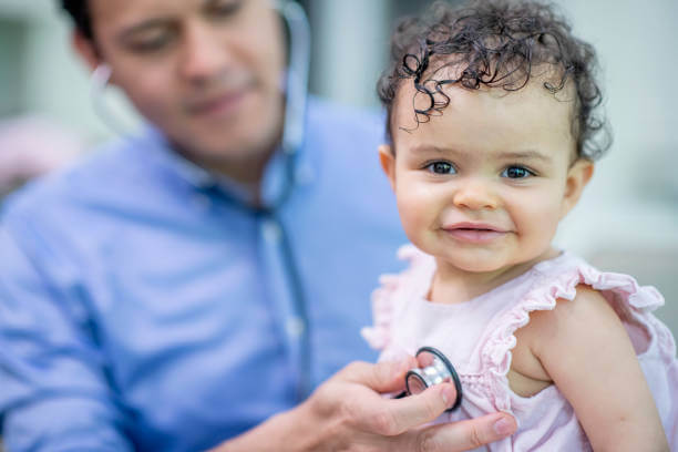 cute toddler is at a medical check up. Her doctor is using a stethoscope to listen to her chest. She is smiling at the camera.